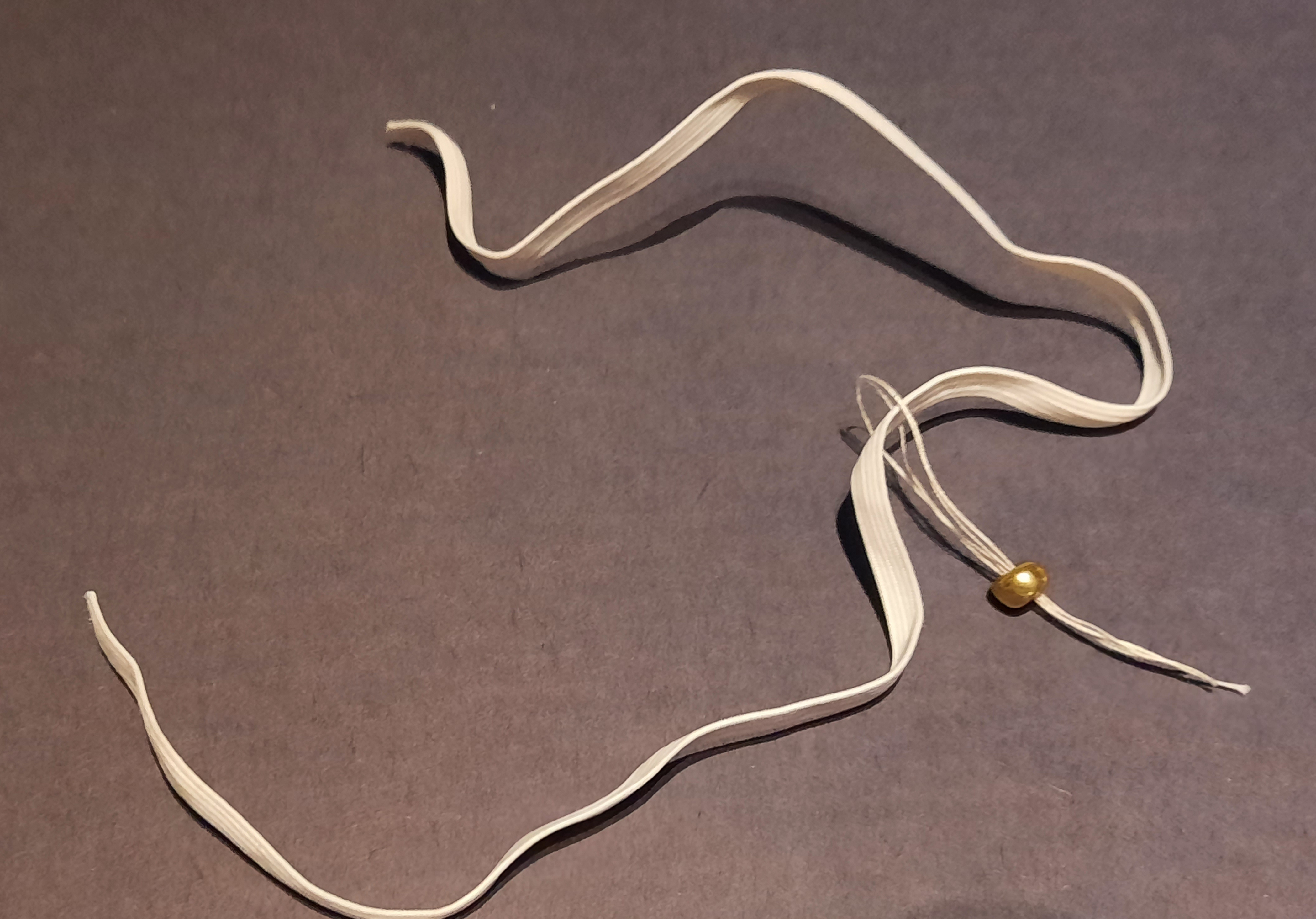 showing how to add a bead to to a piece of elastic intended for a mask - put thread through the bead, over the elastic and back through the bead, then tug on the ends of the threads until the elastic eases out through the bead