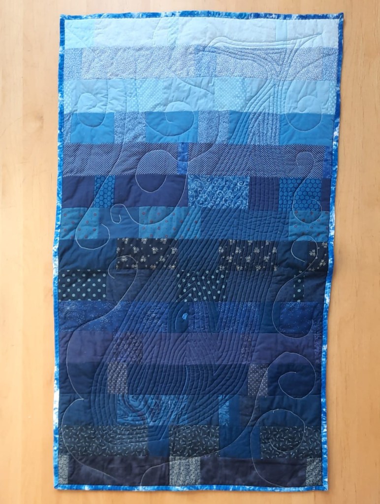 'Now you see her, now you don't' quilted wall-hanging by Amanda Jane Ogden, depicting a diving whale quilted onto a background of blue rectangles